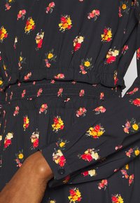 Musta kukkamekko, jossa on pieniä punaisia ja keltaisia kukkakuoseja. Kerrostettu vyötärö joustavalla yksityiskohdalla ja pitkät hihat nappikiinnityksellä.