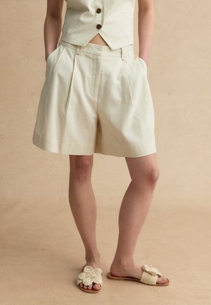 Persoon met beige geplooide shorts, bijpassend gilet met knopen en geweven instapsandalen met grote bloemversiering.