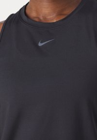 Záběr zblízka na osobu v černém bezrukávovém tričku s malým šedým logem Nike swoosh umístěným ve středu u výstřihu.