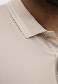 Bež polo majica s teksturiranim materijalom, dugmadi na ovratniku i glatkom završnom obradom s vidljivim detaljima šivanja i suptilnim sjajem.