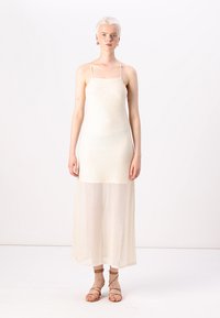 Robe maxi crème, légère, avec des bretelles spaghetti, présentant un motif texturé et un ourlet fluide. Associée à des sandales beiges à lanières.