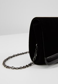 Pochette en velours noir avec bordure noire brillante et bandoulière en chaîne métallique, posée sur une surface blanche devant un fond gris clair.