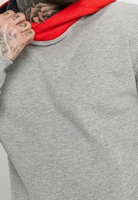 Grå sweatshirt med en röd huva, med en mjuk textur och en ribbad halsringning. Påfallande detalj: intrikata tatueringar synliga på handleden.