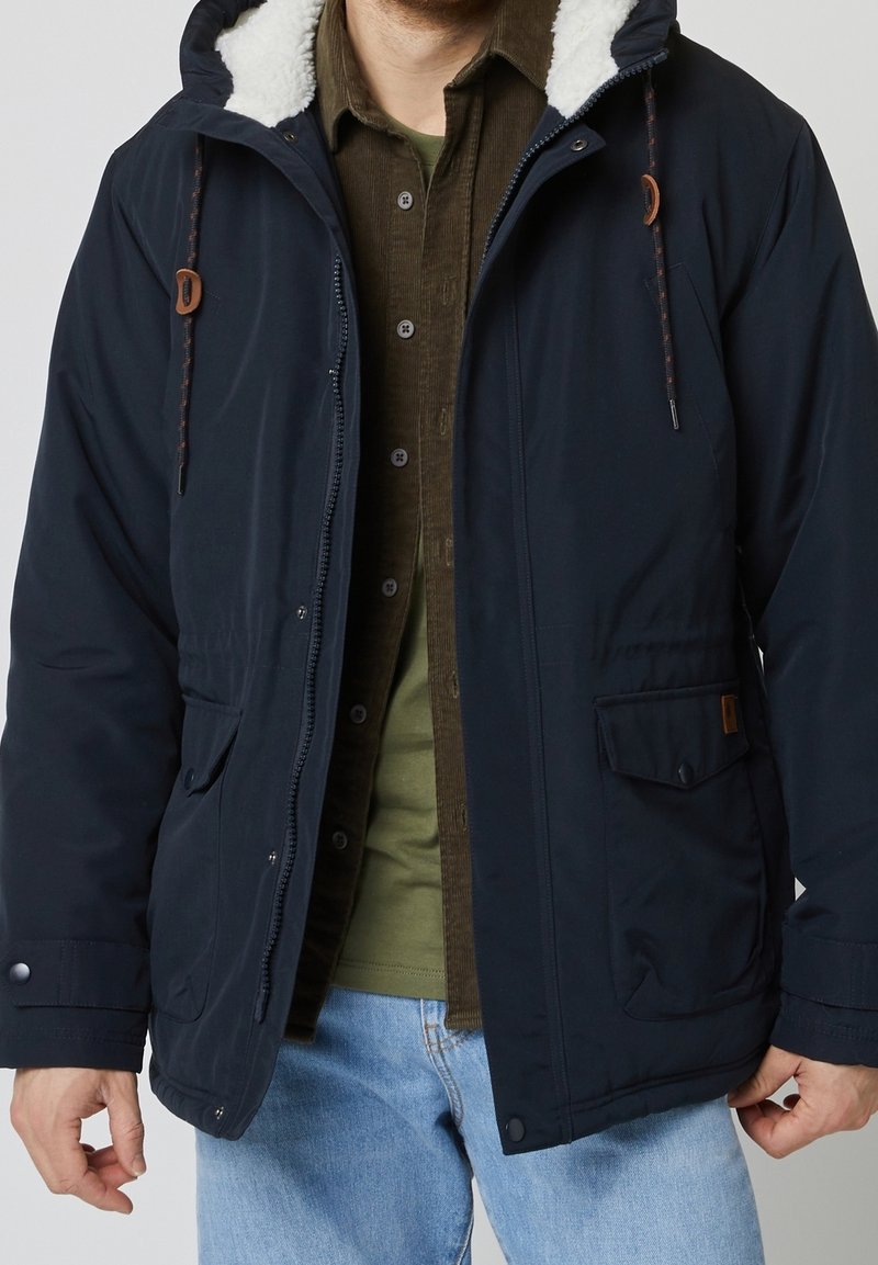 Veste bleu marine avec une capuche doublée en polaire blanche, équipée d'une fermeture éclair, de boutons-pression et de poches latérales. Portée par-dessus une chemise verte.
