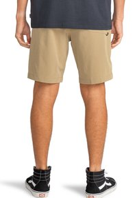 Hombre con pantalones cortos beige y zapatillas altas negras, de pie con las manos relajadas a los lados, de espaldas a la cámara.