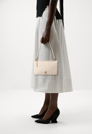 Personne portant une jupe midi blanche et un haut noir, tenant un petit sac à main beige texturé avec une poignée courte, chaussée de chaussures noires à bout pointu.