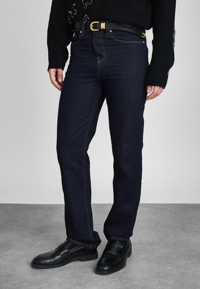 Homme portant un jean bleu foncé, une ceinture en cuir noir avec une boucle dorée, un pull noir et des mocassins en cuir noir, se tenant sur un sol gris.