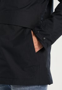 Černá, voděodolná bunda s přední kapsou a knoflíkovým detailem na manžetě, vyrobená z hladké, lehké tkaniny.