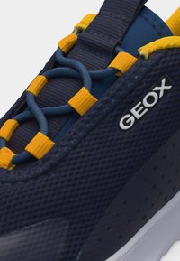 Κοντινό πλάνο ναυτικού μπλε αθλητικού παπουτσιού Geox με κίτρινους βρόχους κορδονιών και ελαστικά κορδόνια bungee σε υφασμάτινο πάνω μέρος με υφή.