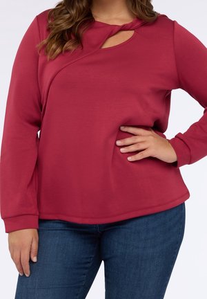 Femme portant un haut rouge à manches longues avec un décolleté en forme de trou de serrure et un jean bleu, main posée sur la hanche.