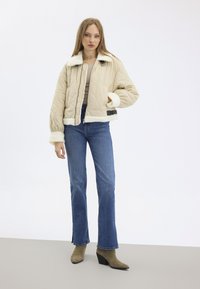 Veste beige matelassée avec col et poignets en fausse fourrure blanche, fermeture éclair, et poches avant plates, associée à un jean bleu évasé et des bottines vertes.