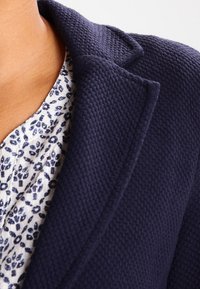 Námořnický strukturovaný blejzr s klopovým límcem, přehozený přes bílé tričko s límcem, které má modrý květinový vzor.