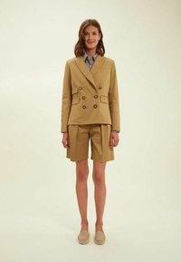 Blazer croisé couleur kaki avec des accents de boutons, associé à un short plissé. Une chemise à col est visible en dessous. Tissu doux et sur mesure.