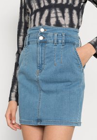 Saia de denim azul com cintura alta, fecho frontal com dois botões, bolsos laterais e um ajuste justo, apresentando costura visível e uma textura suave.