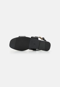 Svart sandal med en strukturerad ovandel, platt gummisula, justerbar rem och spänne i guldton. Har en slät yta och en minimalistisk design.