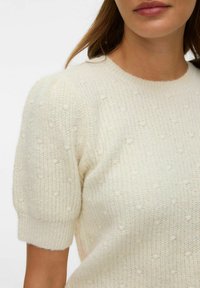 Pull en maille texturée crème avec manches courtes bouffantes, présentant un col côtelé et de petits pois en relief subtil sur tout le tissu.