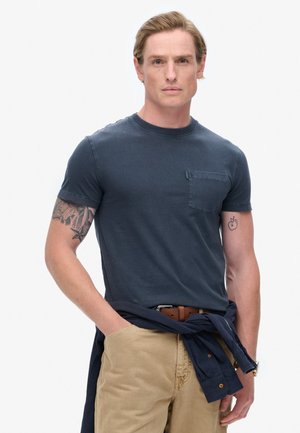 Marineblå crew neck t-shirt lavet af bomuld, med en brystlomme, båret med khaki bukser og en marineblå skjorte bundet om livet.