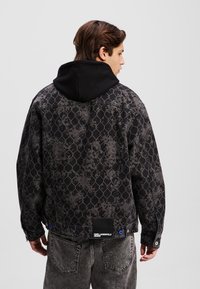 Giacca in denim nera con un design grigio a motivi, dotata di un cappuccio nero, dettagli blu e un'etichetta del marchio visibile all'orlo.