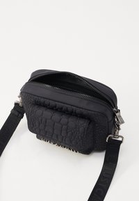 Sac bandoulière noir avec devant en cuir texturé, dos lisse, double zips sur le dessus et bandoulière ajustable avec lettrage de marque.
