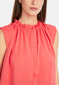 Coral blouse with a ruffled neckline and sleeveless design. Features soft fabric and pleating down the front for added texture.