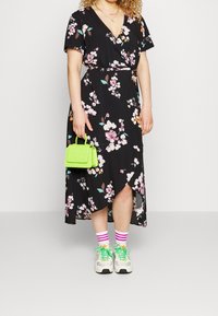 Schwarzes Blumenkleid mit kurzen Ärmeln und einem hohen Schlitz. Kombiniert mit weißen Turnschuhen, pink-weiß gestreiften Socken und einer kleinen grünen Tasche.