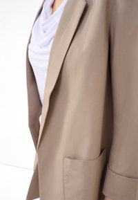 Gros plan sur un blazer beige avec une poche sur un haut blanc drapé, montrant une partie du bras et du torse sur un fond uni.