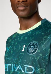 Groen doelmanshirt met driehoekige patronen, een ronde Manchester City-badge, wit nummer 1 en een lichtgroene kraag.
