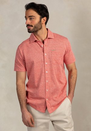 Man met baard, gekleed in een koraalroze overhemd met korte mouwen en lichtbeige broek, kijkt naar links met één hand in zijn broekzak.