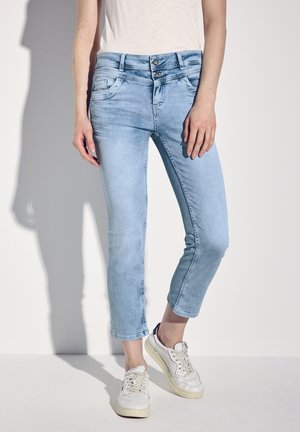 Světle modré vystřižené džíny slim fit s dvoubotovým zapínáním a jemným ošupováním. Kombinované s bílými teniskami. Hladká denimová textura.