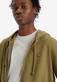Oliven grønn zip-up hettegenser med en grå T-skjorte under. Har et rødt Levi's logo-lapp, snørehette og glatt stofftekstur.