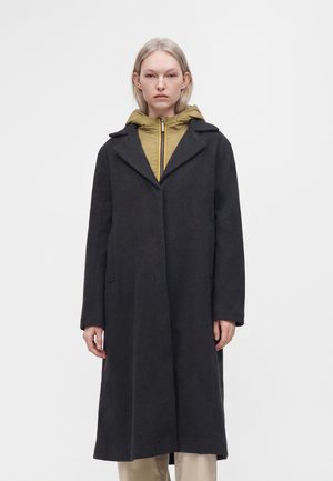 Personne aux cheveux clairs portant un long manteau noir par-dessus une veste à capuche moutarde zippée et un pantalon beige sur un fond clair uni.