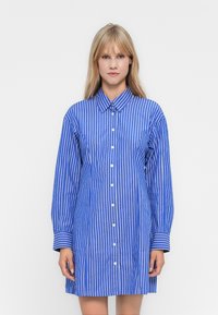 Frau in einem blau-weißen, senkrecht gestreiften Langarmhemdkleid, vor einem schlichten weißen Hintergrund stehend und nach vorne blickend.