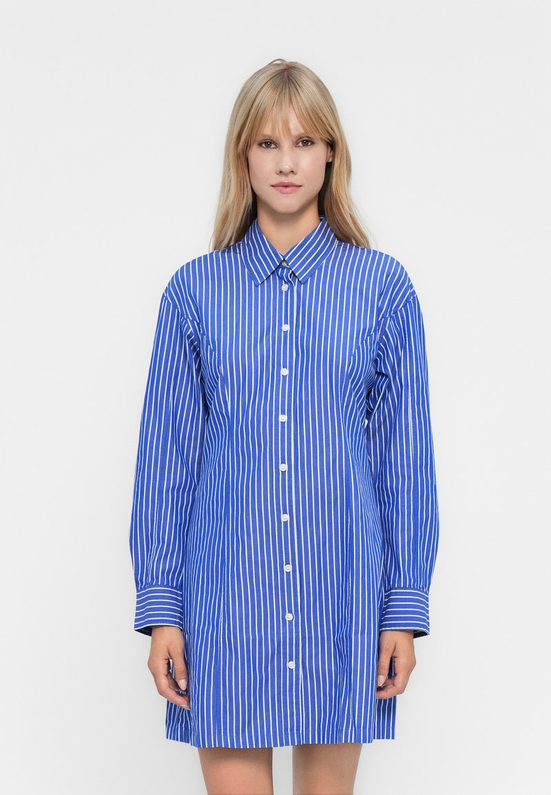 Frau in einem blau-weißen, senkrecht gestreiften Langarmhemdkleid, vor einem schlichten weißen Hintergrund stehend und nach vorne blickend.