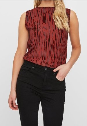 Mouwloze roestrode top met zwarte verticale tijgerstrepen, gedragen met een zwarte high-waist broek, hand rustend in de voorzak.