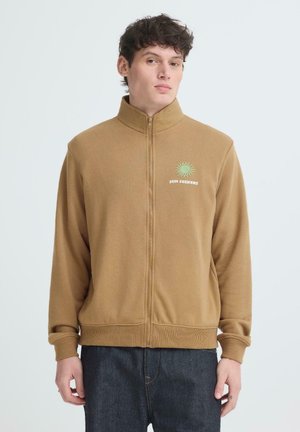 Ung mann iført en lys brun fleecejakke med glidelås, grønt sollogo og teksten "SUN SEEKERS" på brystet, kombinert med mørke jeans.