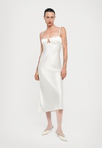 Witte satijnen jurk met een scalloped halslijn, dunne bandjes en een getailleerd silhouet, die tot halverwege de kuit valt. Gepaarde met witte puntige schoenen.