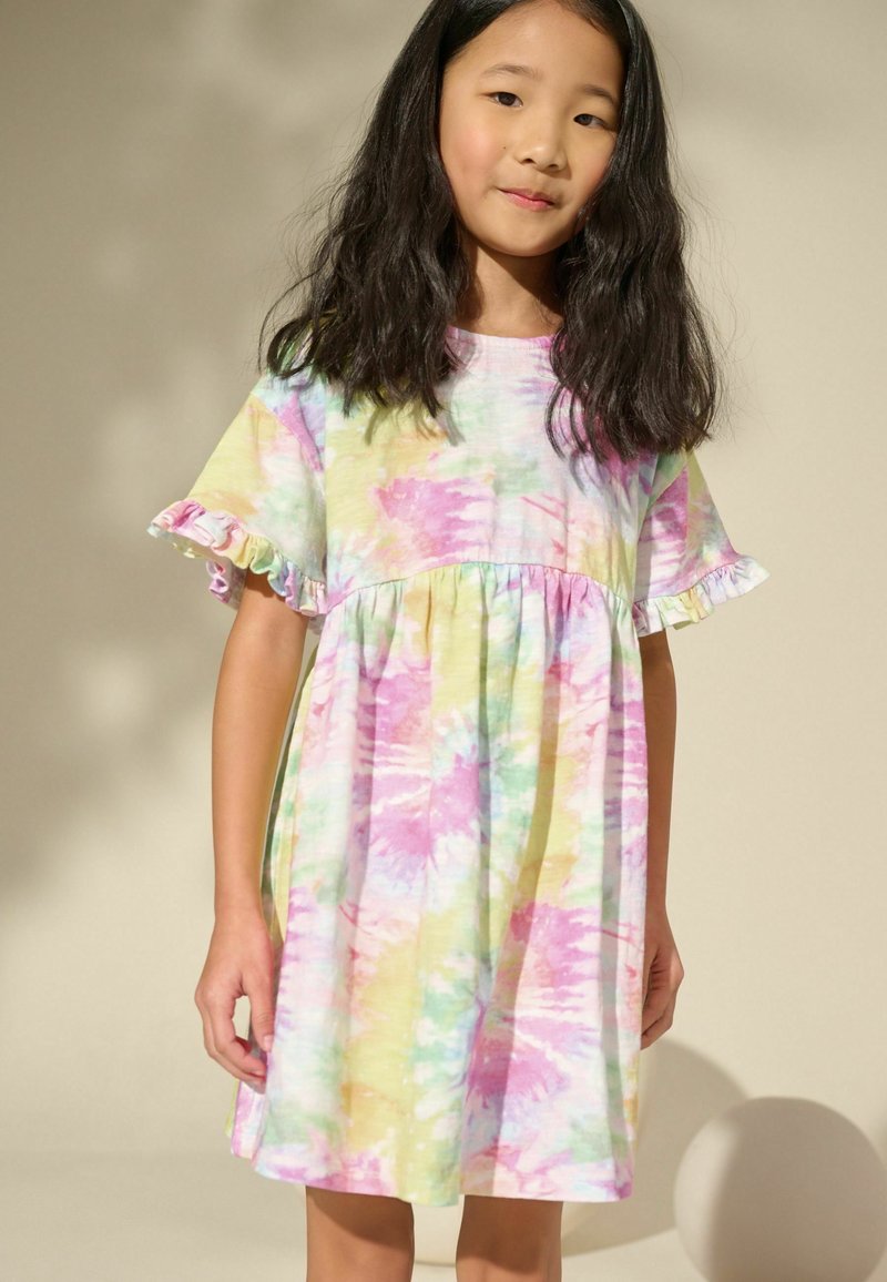 Young girl with long black hair wearing a pastel tie-dye dress with short ruffled sleeves standing against a plain light background.