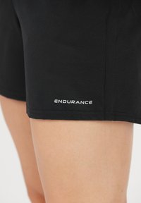 Svarta sportshorts tillverkade av mjukt tyg, med en vit "ENDURANCE"-logotyp vid fållen, med en enkel design och slät textur.