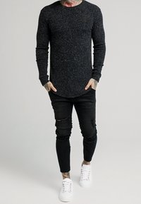 Muž v tmavě šedém svršku s dlouhým rukávem, potrhaných černých skinny džínách a bílých teniskách, stojící s rukama v kapsách.