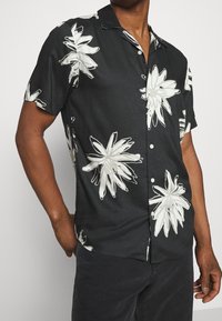 Uomo che indossa una camicia nera a maniche corte con grandi stampe floreali bianche, abbinata a pantaloni scuri, con una mano in tasca.