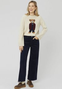 Un pull crème avec un motif d'ours en tricot, assorti à un pantalon en velours côtelé bleu marine à jambes larges. Des chaussures brunes décontractées avec lacets et des chaussettes texturées.