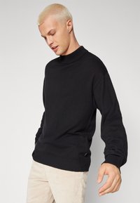 Suéter negro de manga larga y cuello de canalé alto, con un corte holgado y tejido texturizado, combinado con pantalones de pana clara.