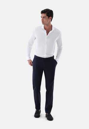 Camicia bianca a maniche lunghe con bottone frontale, abbinata a pantaloni eleganti blu scuro. Scarpe casual completano l'outfit. Design semplice e pulito.