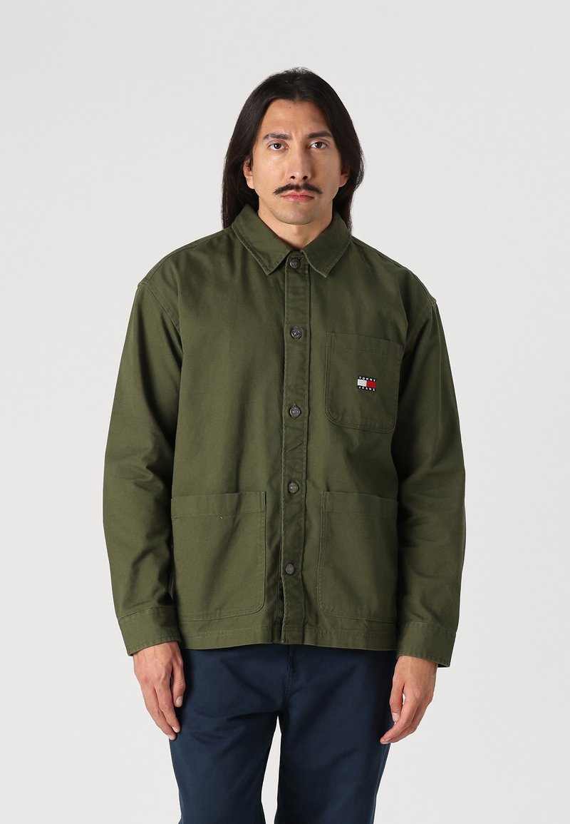 Veste olive, à boutons, avec un col, deux poches avant et une poche poitrine arborant un logo rouge, blanc et bleu. Tissu en mélange de coton.