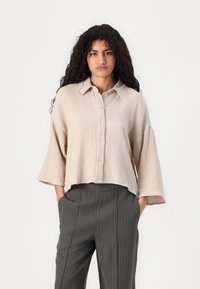 Bežena srajca na gumbe z teksturirano tkanino, ovratnikom in širokimi rokavi, v kombinaciji s sivo prugastimi hlačami.