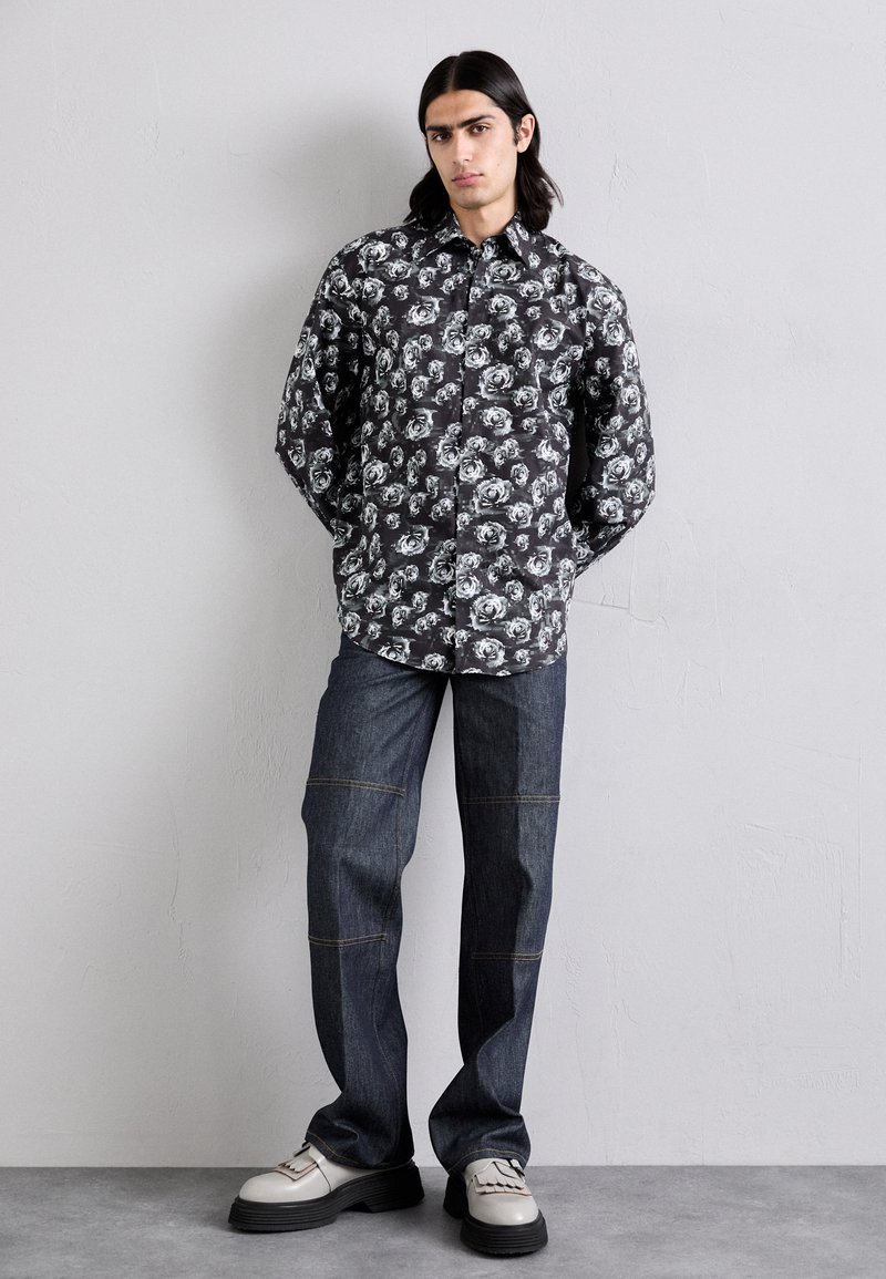 Schwarze Hemd mit Knöpfen und weißem Blumenmuster, kombiniert mit dunkelblauen Jeans, die mit kontrastierenden Nähten und klobigen grauen Schuhen versehen sind.