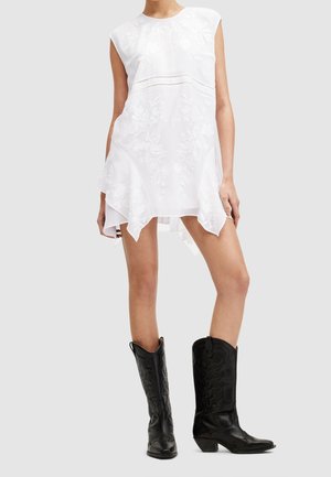 Robe blanche sans manches avec broderie florale et ourlet asymétrique, associée à des bottes de cowboy noires mi-mollet avec coutures décoratives.