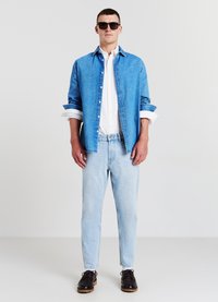 Camicia di denim azzurro sopra una camicia bianca con bottoni, abbinata a jeans azzurri e scarpe stringate scure. Gli occhiali da sole neri completano il look.