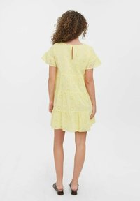 Geel gelaagd jurkje met bloemmotief in ajour, korte zwierige mouwen en een keyhole-detail aan de achterkant. De zoom valt boven halverwege het dijbeen.