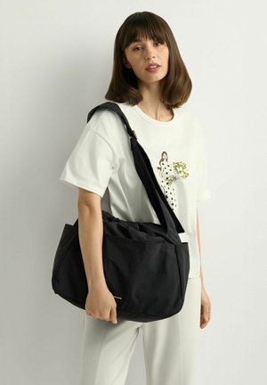 Kobieta o średniej długości brązowych włosach nosi białą koszulkę z nadrukiem w białe kropki i dużą czarną torbę na ramię na jasnym tle.